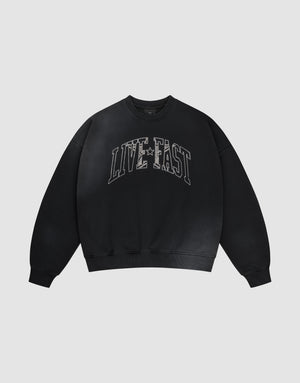 The LIVE FAST DIE YOUNG Live Fast Rhinestone Sweater is a black oversized crewneck with bold distressed white "LIVE FAST" lettering and a subtle spray effect, set against a plain light gray background. | plain