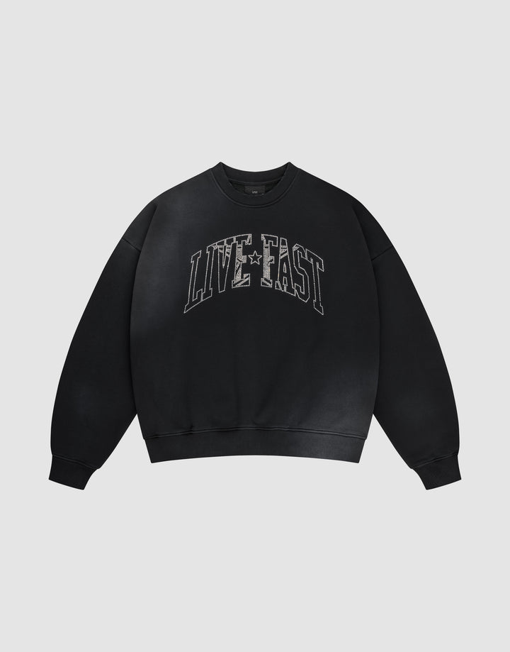 The LIVE FAST DIE YOUNG Live Fast Rhinestone Sweater is a black oversized crewneck with bold distressed white "LIVE FAST" lettering and a subtle spray effect, set against a plain light gray background. | plain