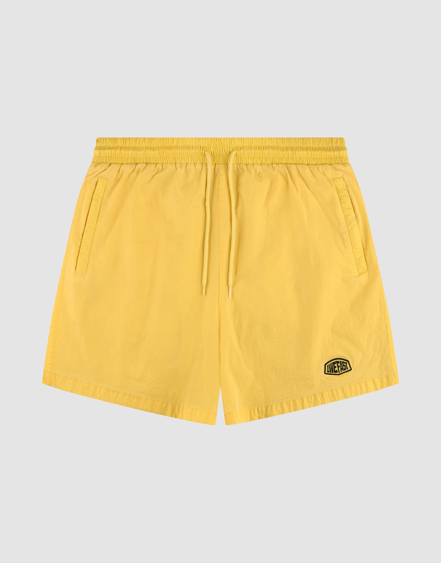 The Live Fast Swim Shorts by LIVE FAST DIE YOUNG feature a regular fit, elastic waistband, drawstring, side pockets, and a small black logo patch on the left leg. Shown in Tinted Yellow Wash laid flat on a light gray background.