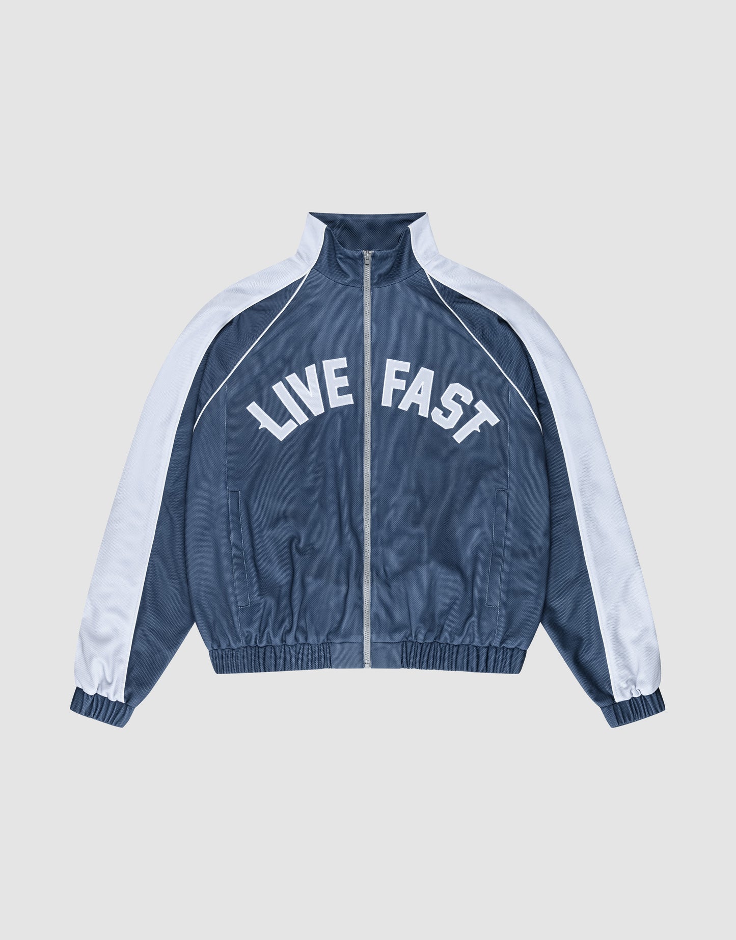The LFDY Arch Trackjacket by LIVE FAST DIE YOUNG is a blue and white zip-up jacket with an oversize fit, featuring "LIVE FAST" in bold white letters across the chest and seitliche Eingrifftaschen for convenience.