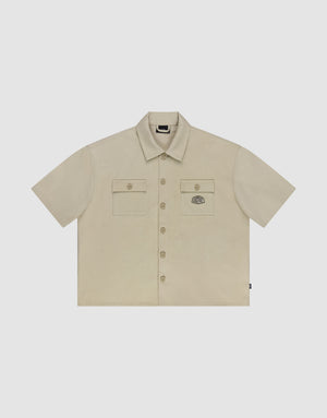 The LFDY Twill Shirt by LIVE FAST DIE YOUNG features a beige cropped oversize fit, 100% cotton, short sleeves, two button-flap chest pockets with an embroidered patch on the left, and a classic collar—shown flat on a white background. | plain