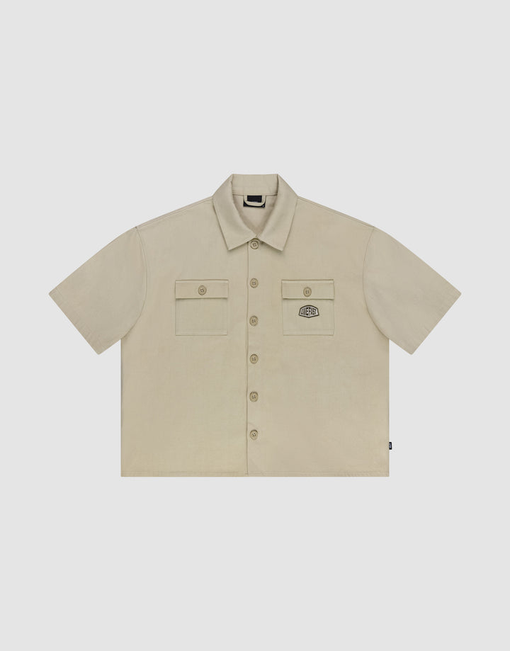 The LFDY Twill Shirt by LIVE FAST DIE YOUNG features a beige cropped oversize fit, 100% cotton, short sleeves, two button-flap chest pockets with an embroidered patch on the left, and a classic collar—shown flat on a white background. | plain