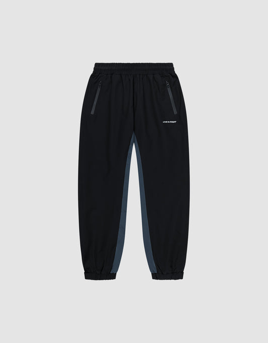 Black Basic Shell Pants by LIVE FAST DIE YOUNG with elastic waistband, zippered side pockets, gray panels, and elastic cuffs. Features a white reflective logo on the left thigh. Displayed on a plain light background. | plain