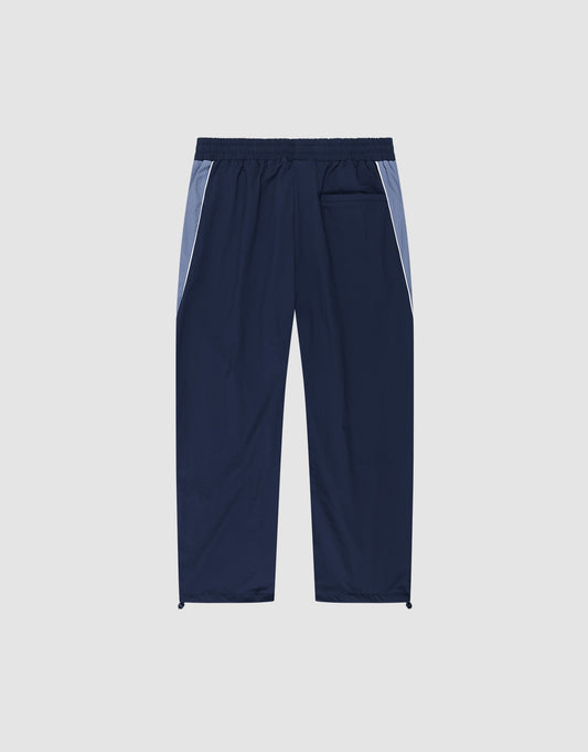 The LIVE FAST DIE YOUNG Basic Nylon Trackpants in navy feature light blue side panels, an Elasticbund waistband, a back pocket, and adjustable drawstring hems. Relaxed Wide Fit silhouette shown against a plain light background.