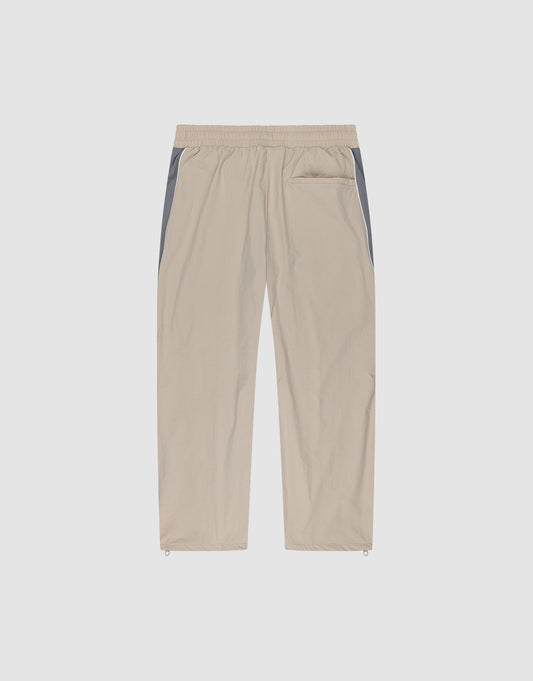 The LIVE FAST DIE YOUNG Basic Nylon Trackpants feature a wide fit, elastic waistband, straight beige legs with grey side panels, single back pocket, and adjustable ankle drawstrings—shown against a plain white background.