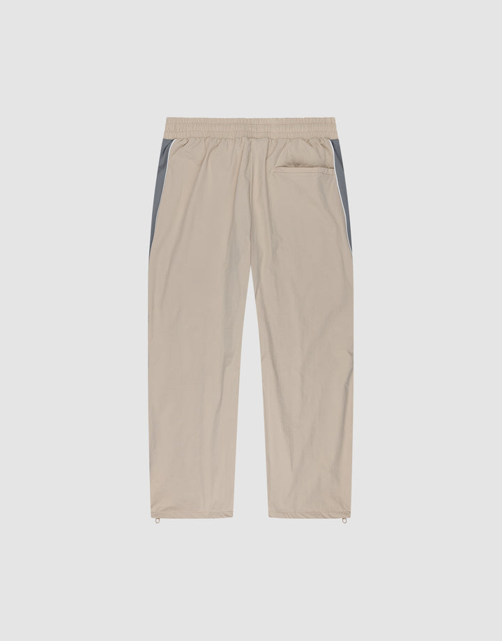 The LIVE FAST DIE YOUNG Basic Nylon Trackpants feature a wide fit, elastic waistband, straight beige legs with grey side panels, single back pocket, and adjustable ankle drawstrings—shown against a plain white background.