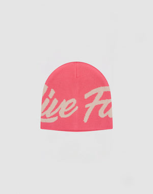 The LFDY Jacquard Beanie by LIVE FAST DIE YOUNG is a pink polyacryl hat with a large cream cursive logo wrapping around it, displayed on a plain light gray background.