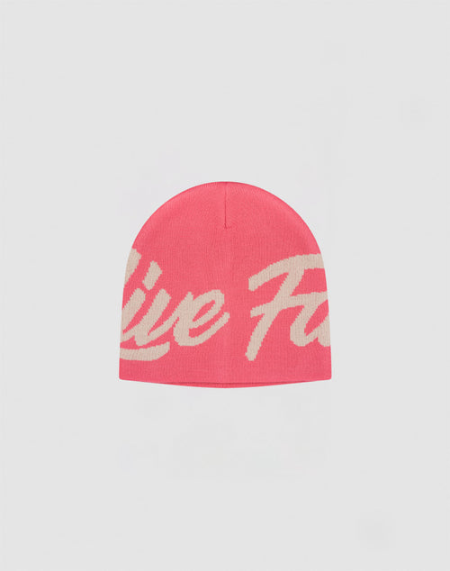 The LFDY Jacquard Beanie by LIVE FAST DIE YOUNG is a pink polyacryl hat with a large cream cursive logo wrapping around it, displayed on a plain light gray background.