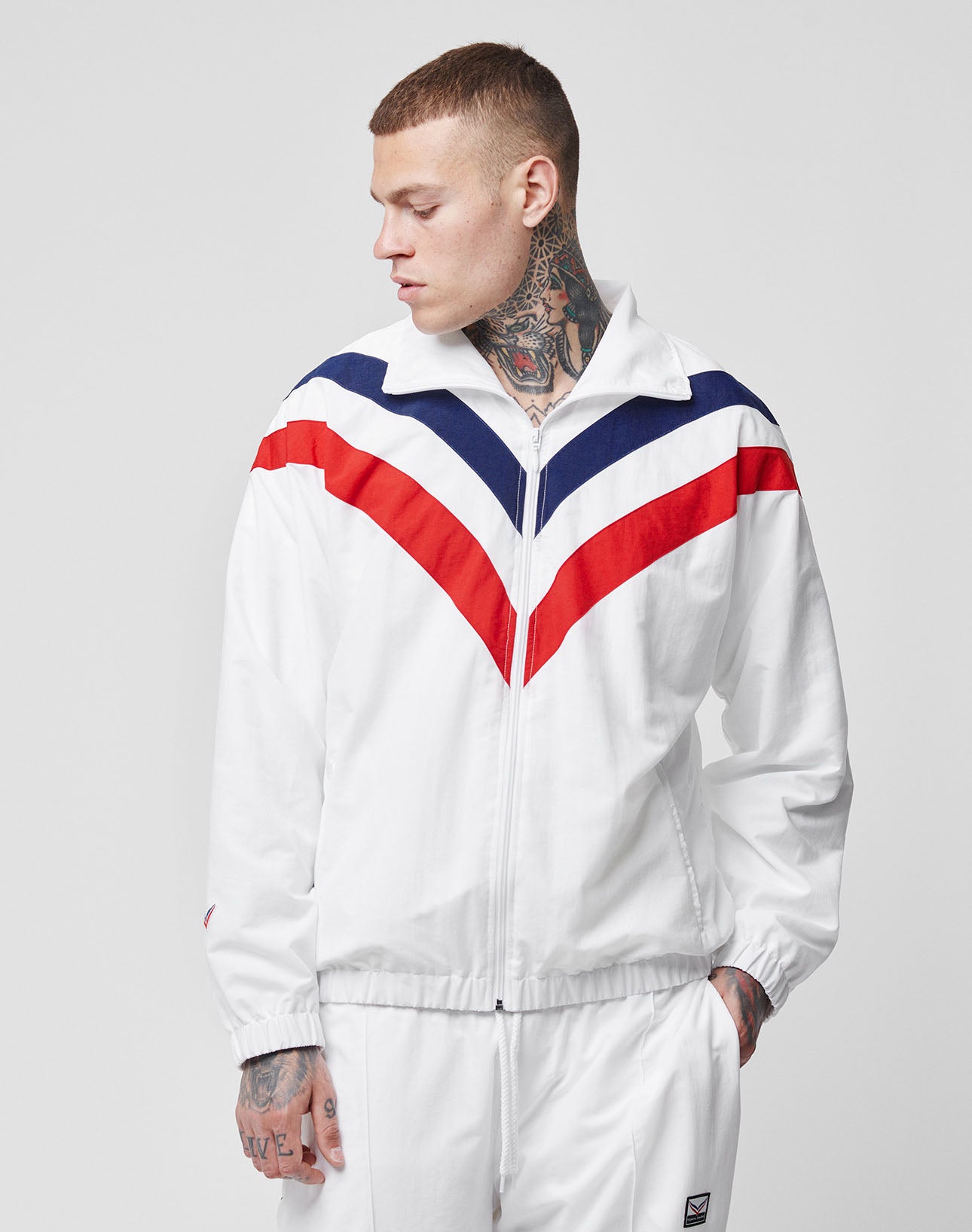 A tattooed man wears the LFDY x Trigema Trackjacket by LIVE FAST DIE YOUNG—a white zip-up with red and blue chevron stripes and Raglan sleeves—looking left with one hand in his pocket against a plain gray background.