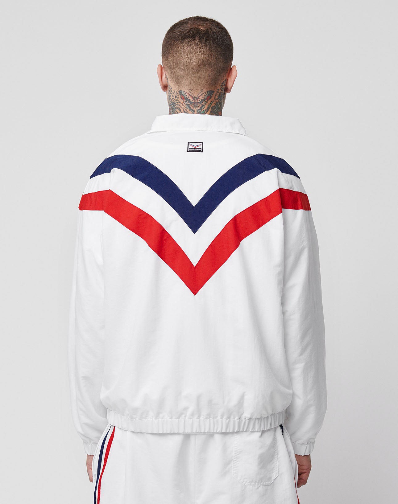 Seen from behind, a person with short hair and neck tattoos wears the LFDY x Trigema Trackjacket by LIVE FAST DIE YOUNG—white with Raglan sleeves, red and navy chevron stripes on the back, plus matching white side-striped pants.