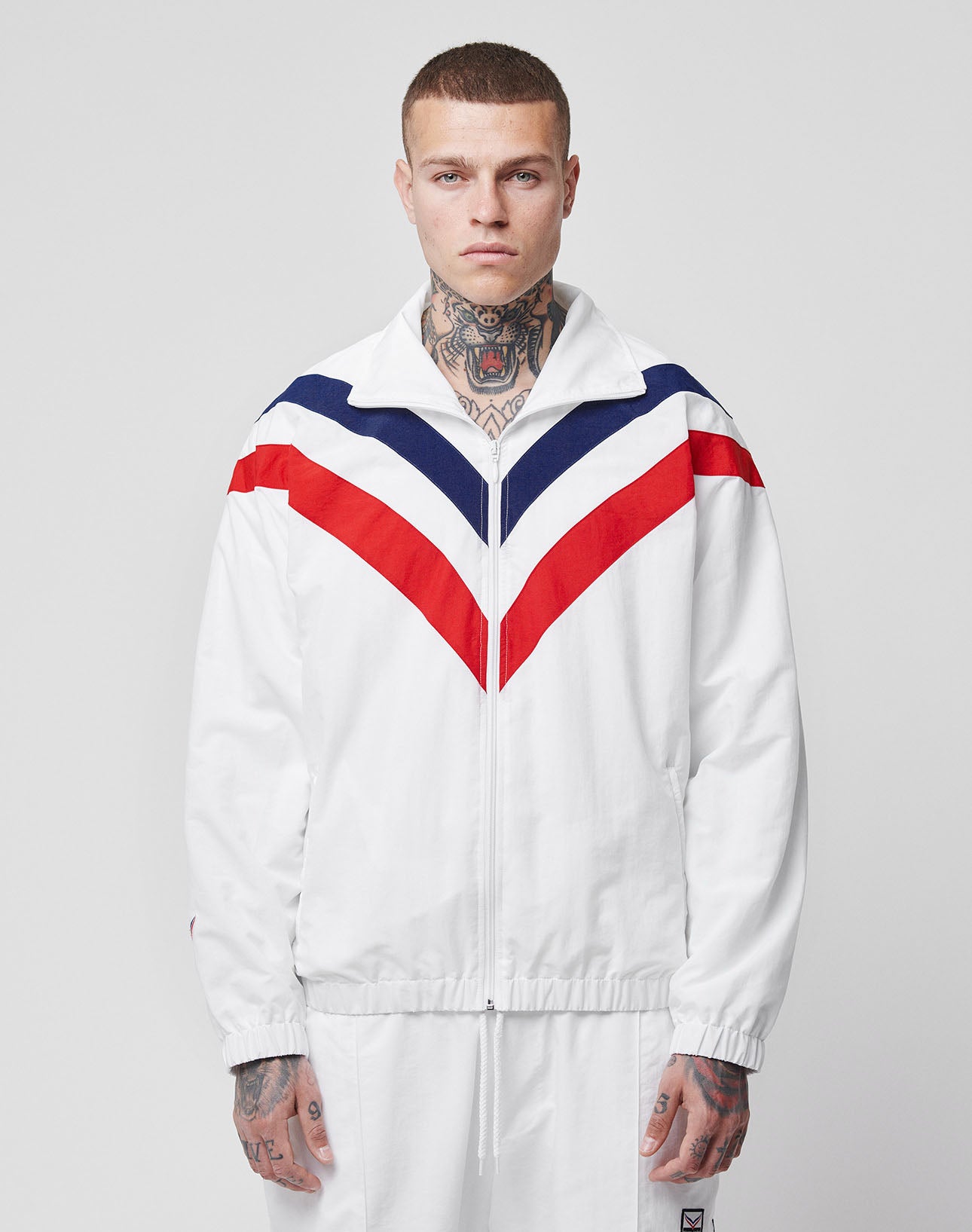 A person with neck and hand tattoos stands facing forward in the limited LFDY x Trigema Trackjacket by LIVE FAST DIE YOUNG, featuring red and navy chevron stripes and matching white pants, against a plain background.
