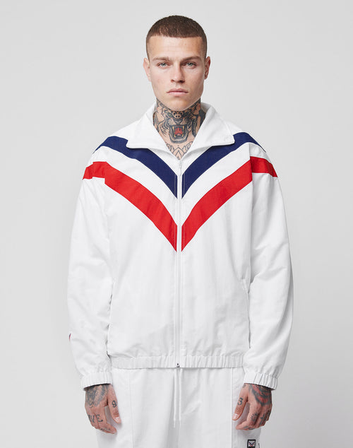 A person with neck and hand tattoos stands facing forward in the limited LFDY x Trigema Trackjacket by LIVE FAST DIE YOUNG, featuring red and navy chevron stripes and matching white pants, against a plain background.