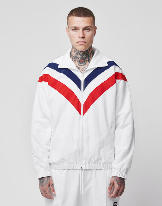 A person with neck and hand tattoos stands facing forward in the limited LFDY x Trigema Trackjacket by LIVE FAST DIE YOUNG, featuring red and navy chevron stripes and matching white pants, against a plain background.