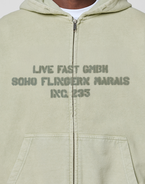 A close-up of the LFDY District Hooded Zip by LIVE FAST DIE YOUNG in light green, featuring a bold front print "LIVE FAST GMBH SOHO FLINGERN MARAIS INC. 235" on the chest. Oversize fit, crafted from 100% Baumwolle (cotton).