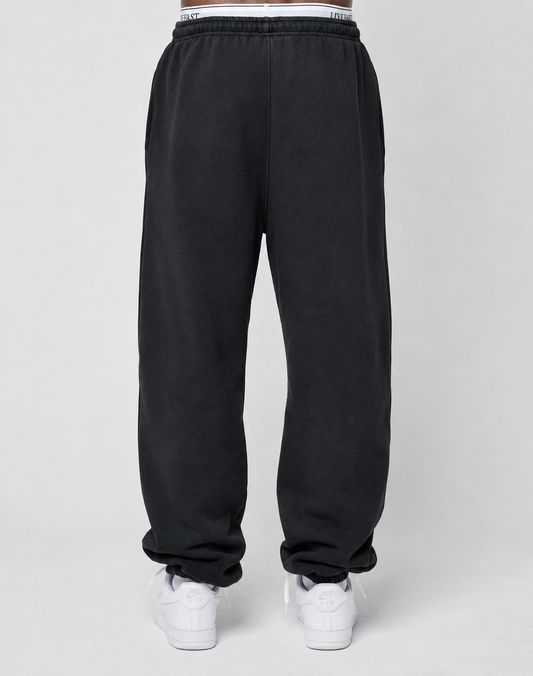 A person models the LIVE FAST DIE YOUNG Basic Loose Jogger in black, featuring an elastic waistband and subtle logo print, paired with white sneakers. The shot is from the back, focusing on the lower body against a plain background.