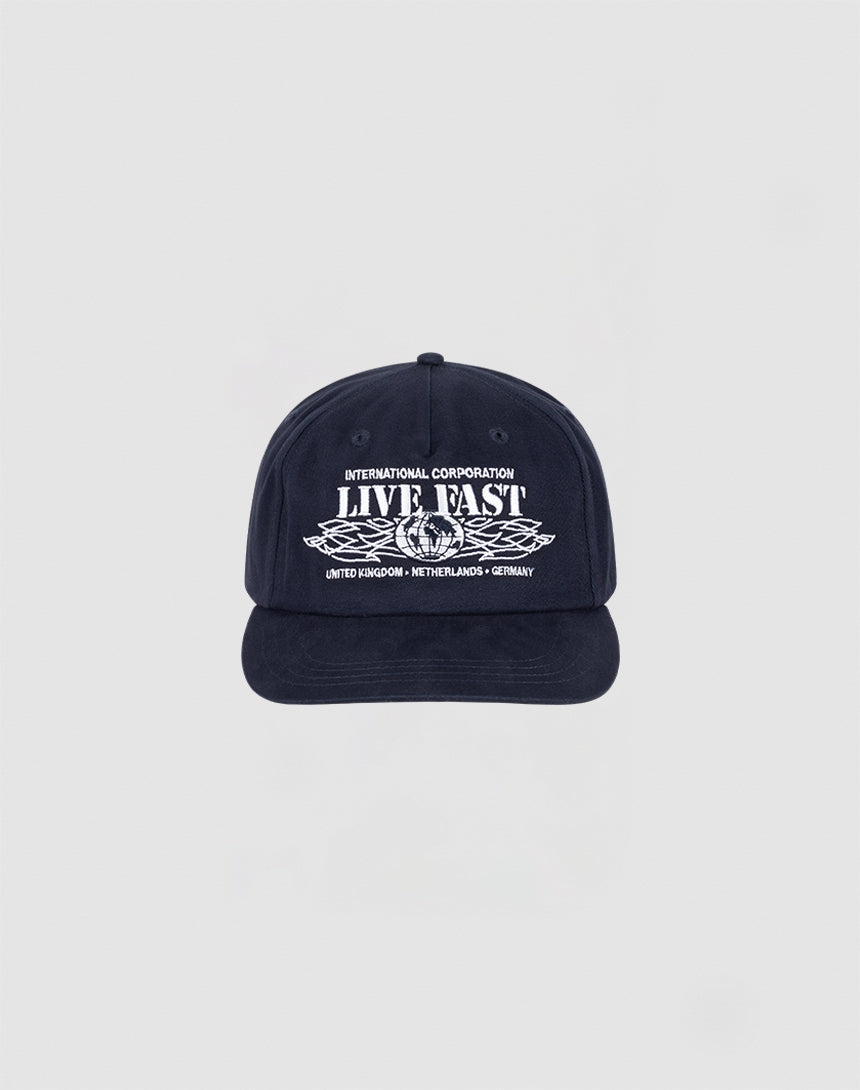 The Live Fast Corporation Cap by LIVE FAST DIE YOUNG is a navy unstructured 5-panel hat with INTERNATIONAL CORPORATION LIVE FAST in white, leaf graphic, multi-logo accents, and a flag label listing UNITED KINGDOM, NETHERLANDS, and GERMANY. 