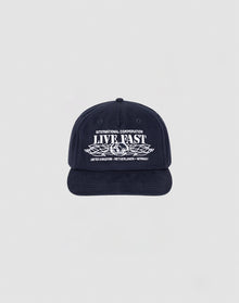 The Live Fast Corporation Cap by LIVE FAST DIE YOUNG is a navy unstructured 5-panel hat with INTERNATIONAL CORPORATION LIVE FAST in white, leaf graphic, multi-logo accents, and a flag label listing UNITED KINGDOM, NETHERLANDS, and GERMANY.