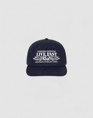 The Live Fast Corporation Cap by LIVE FAST DIE YOUNG is a navy unstructured 5-panel hat with INTERNATIONAL CORPORATION LIVE FAST in white, leaf graphic, multi-logo accents, and a flag label listing UNITED KINGDOM, NETHERLANDS, and GERMANY.