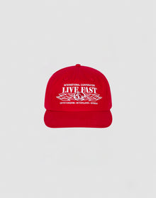 The Live Fast Corporation Cap by LIVE FAST DIE YOUNG is a 100% cotton red cap with bold white International Corporation LIVE FAST text and multi-logo graphics, plus country names United Kingdom, Netherlands, and Germany on the front.