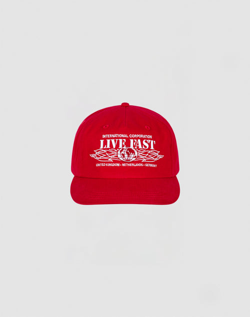 The Live Fast Corporation Cap by LIVE FAST DIE YOUNG is a 100% cotton red cap with bold white International Corporation LIVE FAST text and multi-logo graphics, plus country names United Kingdom, Netherlands, and Germany on the front.