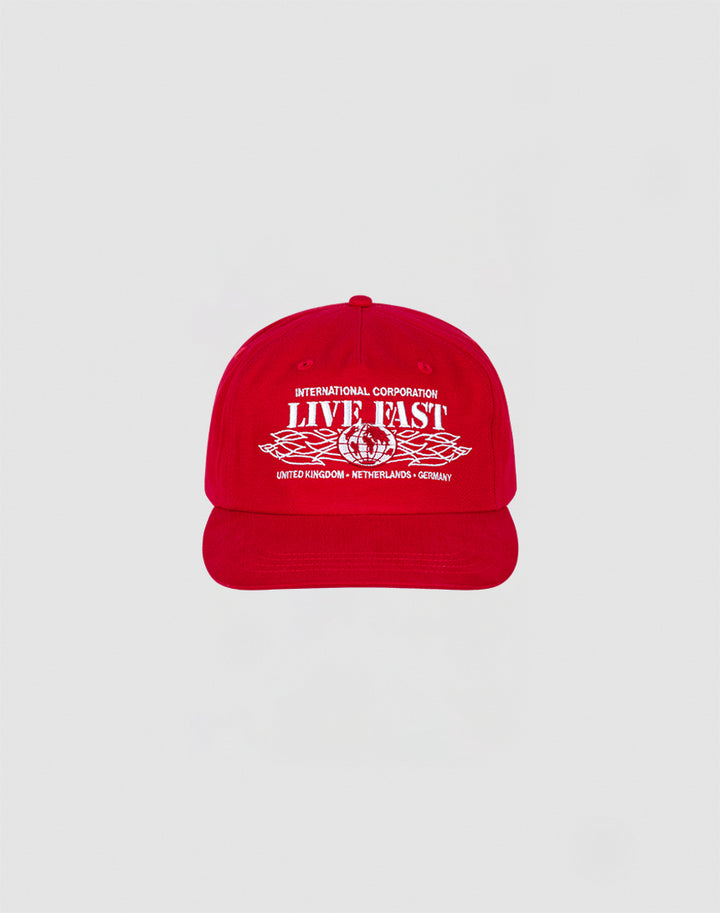 The Live Fast Corporation Cap by LIVE FAST DIE YOUNG is a 100% cotton red cap with bold white International Corporation LIVE FAST text and multi-logo graphics, plus country names United Kingdom, Netherlands, and Germany on the front.