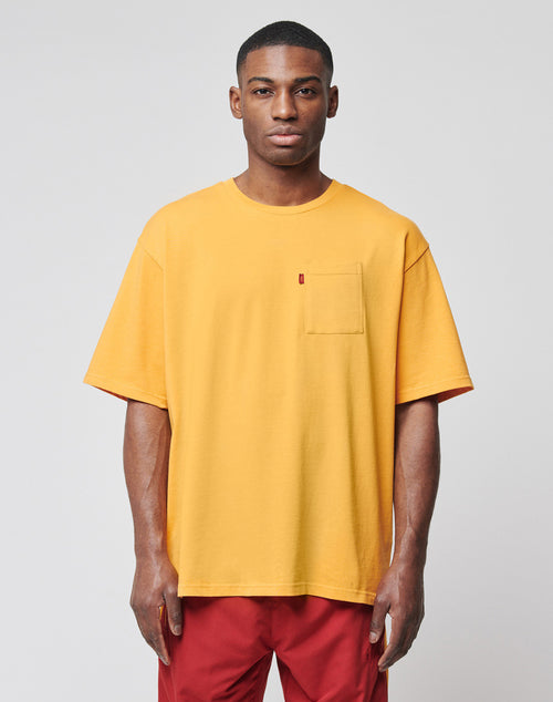 A man faces the camera with a neutral expression, wearing a LIVE FAST DIE YOUNG LFDY Tube Pocket Tee in yellow with a left chest pocket and red pants against a plain gray background.