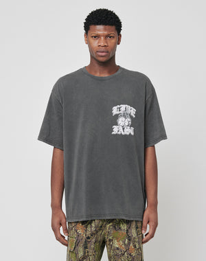 A man faces forward wearing the LIVE FAST DIE YOUNG Burning Dice Tee, a regular fit faded black cotton T-shirt with a white graphic and text on the left chest, paired with camouflage pants against a plain light gray background.