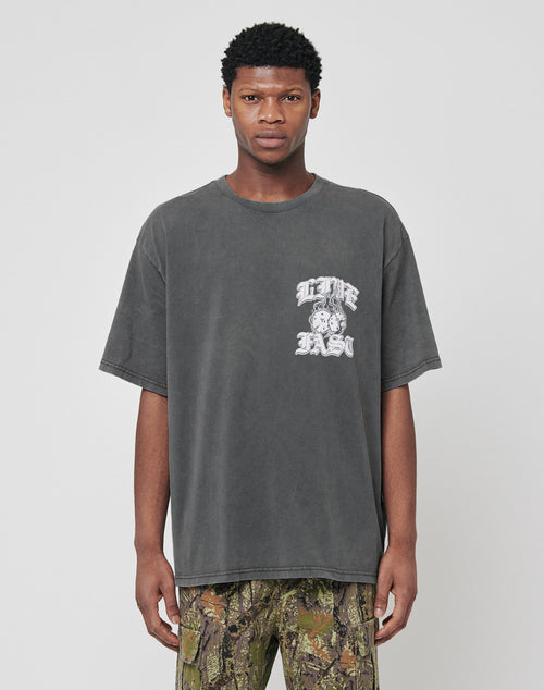 A man faces forward wearing the LIVE FAST DIE YOUNG Burning Dice Tee, a regular fit faded black cotton T-shirt with a white graphic and text on the left chest, paired with camouflage pants against a plain light gray background.