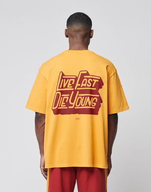 Someone stands facing away from the camera against a plain background, wearing the LIVE FAST DIE YOUNG LFDY Tube Pocket Tee in yellow with Live Fast Die Young boldly printed on the back, paired with matching pants.