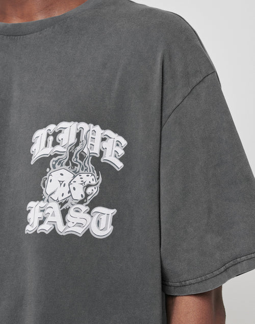 Someone wears the Burning Dice Tee by LIVE FAST DIE YOUNG, a regular fit cotton shirt in faded gray with bold gothic white LIVE FAST lettering and flaming dice artwork printed on the chest.