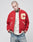 A person with short hair and neck tattoos wears the red LFDY OC College Jacket with cream trim, a yellow C patch, and Organized Crime text over a white shirt and blue jeans, standing hands in pockets against a plain background.