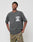 A person wearing a regular fit, dark gray Burning Dice Tee by LIVE FAST DIE YOUNG with white gothic LIVE FAST lettering and chest graphic, paired with camouflage cargo pants, stands hands in pockets against a plain background.