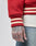 A close-up of a tattooed hand with LIVE on the fingers, wearing LFDYs OC College Jacket in red with cream/red striped cuffs, Rücken-Stick Frottee-Patch detail, Eingrifftaschen, a white shirt, and blue jeans.