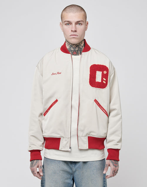 A person with neck and hand tattoos wears the cream-colored LFDY OC College Jacket, featuring red trim, red pockets, and a large red terry O patch on the chest, standing against a plain light background.