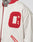 Close-up of a person wearing the cream-colored OC College Jacket by LFDY with red trim and a large red-white terry O patch on the chest; part of their neck tattoo is visible.