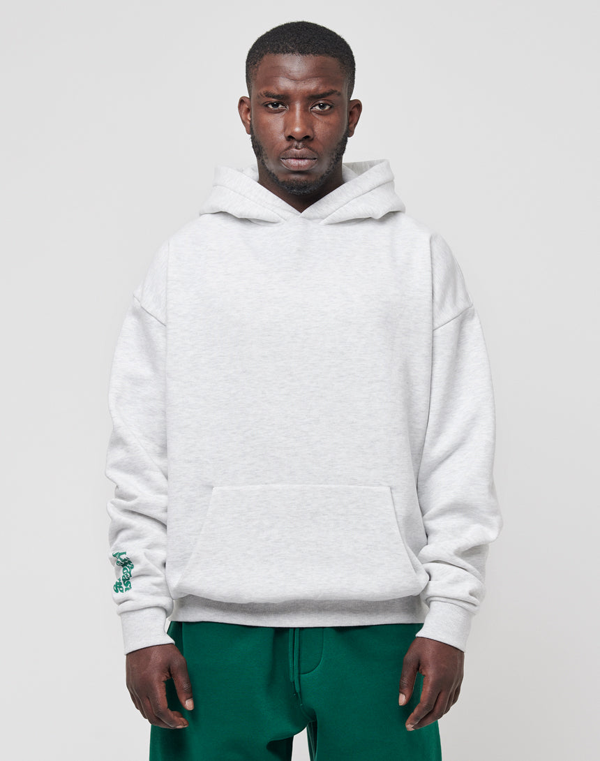 A man wears a light gray LIVE FAST DIE YOUNG Basic Wrist Hooded sweatshirt with a front pocket and green pants, standing against a plain light gray background for a subtle vintage effect.