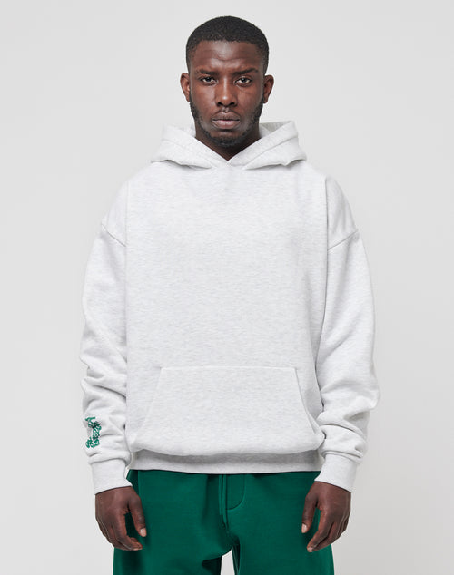 A man wears a light gray LIVE FAST DIE YOUNG Basic Wrist Hooded sweatshirt with a front pocket and green pants, standing against a plain light gray background for a subtle vintage effect.