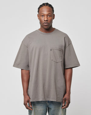 A man with braided hair wears the LFDY Destroyed Pocket Tee in gray, featuring a chest pocket and subtle distressed details. He pairs it with light blue jeans and stands against a plain light gray background.