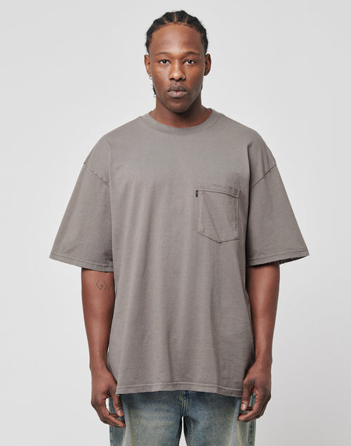 A man with braided hair wears the LFDY Destroyed Pocket Tee in gray, featuring a chest pocket and subtle distressed details. He pairs it with light blue jeans and stands against a plain light gray background.