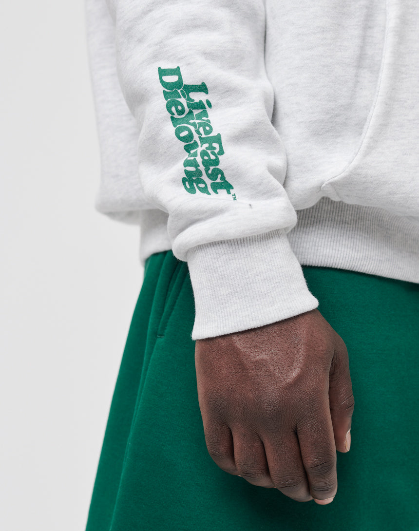A person wears the LIVE FAST DIE YOUNG Basic Wrist Hooded sweatshirt in light gray with green sleeve text, paired with green pants. Visible from the waist down, their hand is relaxed by their side. Subtle vintage effect adds a cool vibe.