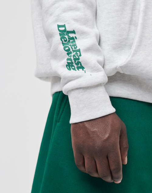 A person wears the LIVE FAST DIE YOUNG Basic Wrist Hooded sweatshirt in light gray with green sleeve text, paired with green pants. Visible from the waist down, their hand is relaxed by their side. Subtle vintage effect adds a cool vibe.