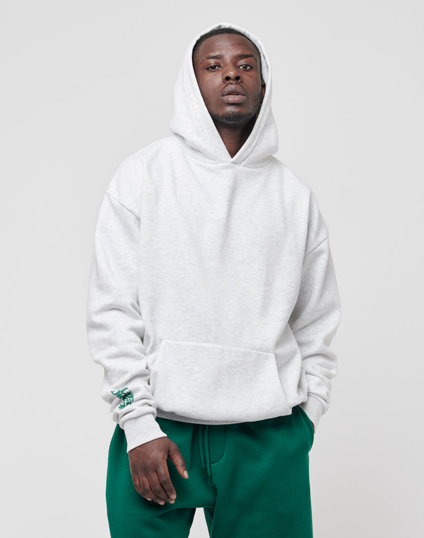 A person wearing a light gray LIVE FAST DIE YOUNG Basic Wrist Hooded with the hood up and green pants stands against a plain, light gray background, looking directly at the camera with hands in the hoodie pocket.