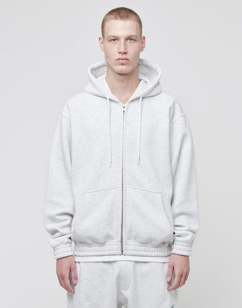 A person wearing the LIVE FAST DIE YOUNG Basic Hooded Zip in light gray, with topstitching and hands in pockets, stands against a plain light background, looking directly at the camera.