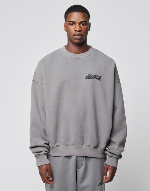 A man faces forward in the LIVE FAST DIE YOUNG Basic Chest Sweater, a gray oversized sweatshirt and sweatpants set with topstitching and small black text on the left chest and thigh, made from a cotton-polyester blend, against a plain light gray background.