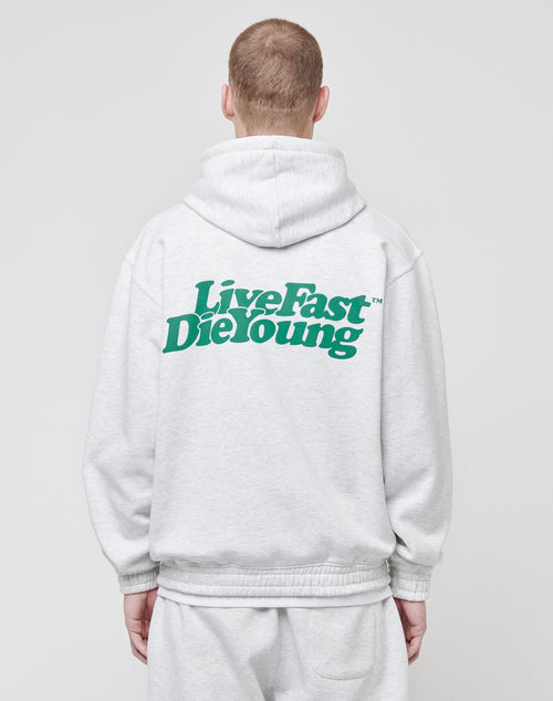 A person is seen from the back wearing the LIVE FAST DIE YOUNG Basic Hooded Zip in light gray with topstitching and matching sweatpants. The hoodie features a bold green Live Fast Die Young print on the back.