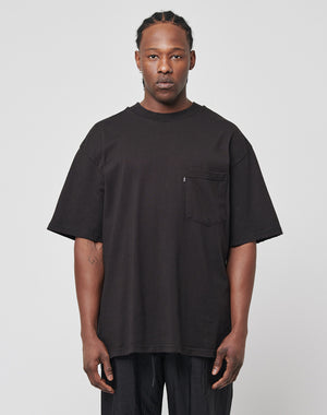 A man with braided hair and a neutral expression wears the LFDY Destroyed Pocket Tee in an oversized black fit with a chest pocket, paired with black pants, standing against a plain light gray background.
