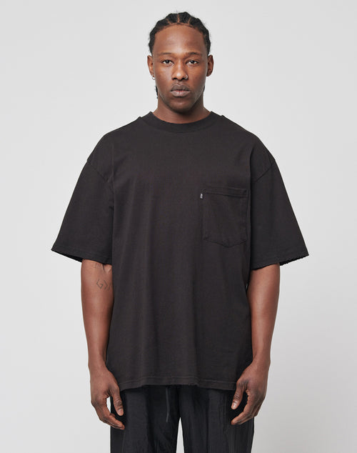 A man with braided hair and a neutral expression wears the LFDY Destroyed Pocket Tee in an oversized black fit with a chest pocket, paired with black pants, standing against a plain light gray background.