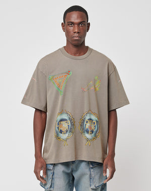 A man wears the LIVE FAST DIE YOUNG Died for It Tee, an oversized, taupe 100% cotton t-shirt with colorful abstract graphics and LIVED FOR IT and DIED FOR IT on the front, paired with blue jeans against a plain white background.