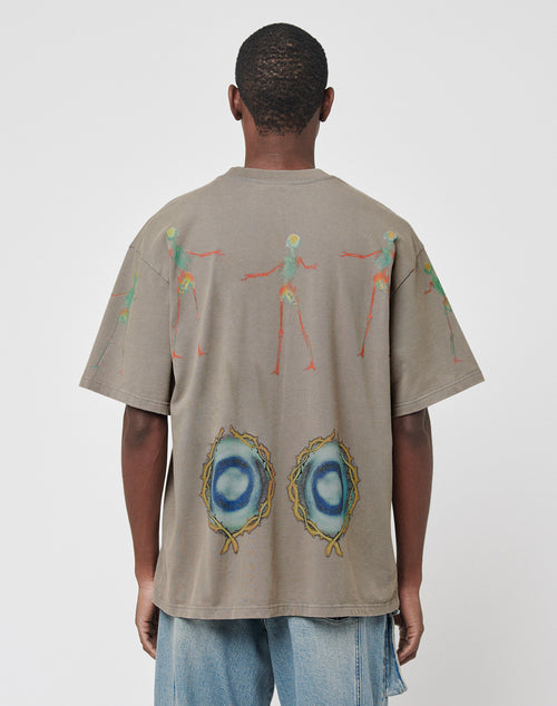 A person facing away wears the LIVE FAST DIE YOUNG Died for It Tee, a gray oversize vintage-effect T-shirt featuring colorful abstract human figures on the upper back and two circular blue/yellow designs near the hem, paired with light blue jeans.