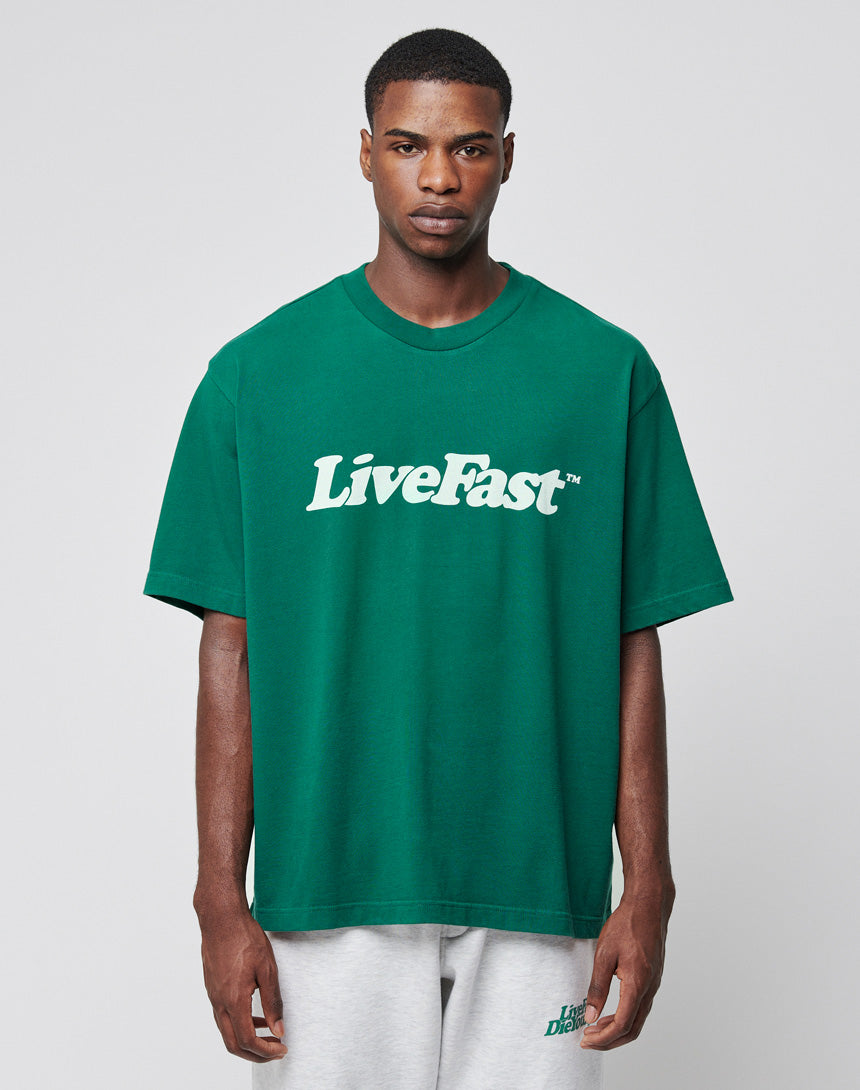 A man stands facing forward against a plain light gray background, wearing the LIVE FAST DIE YOUNG Basic Tee in green with a white LiveFast print and light gray 100% Baumwolle shorts. He has a serious expression.